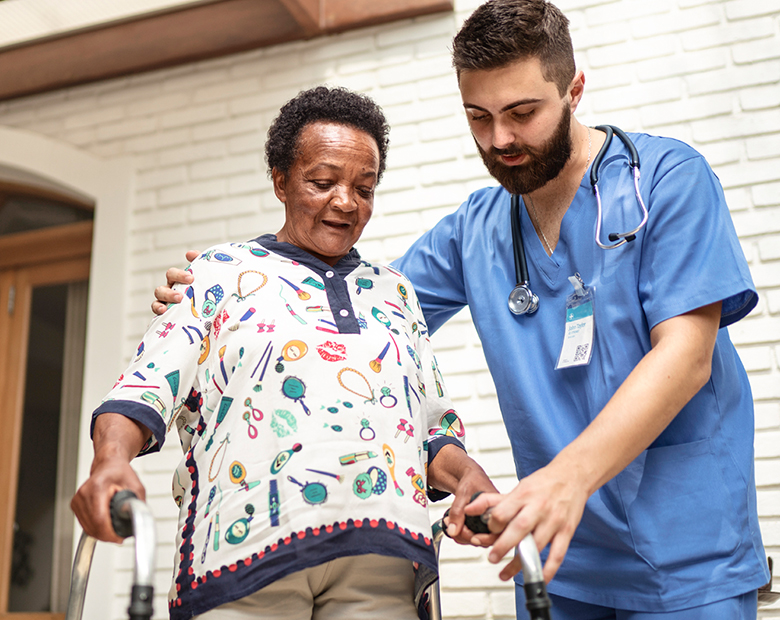 A healthcare worker supports an older adult using a walker outside a home, showing care, mobility support, and trust.